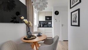 A modern dining area with a round wooden table, two grey chairs, and a vase with flowers. A kitchen is visible in the background with white cabinets and a microwave. - Petit Repit by Grand Apartments (Sopot)
