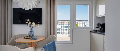 Una sala de estar moderna con una mesa redonda de madera, dos sillas, un jarrón azul con flores, una lámpara de araña blanca grande y un balcón con vista a edificios.