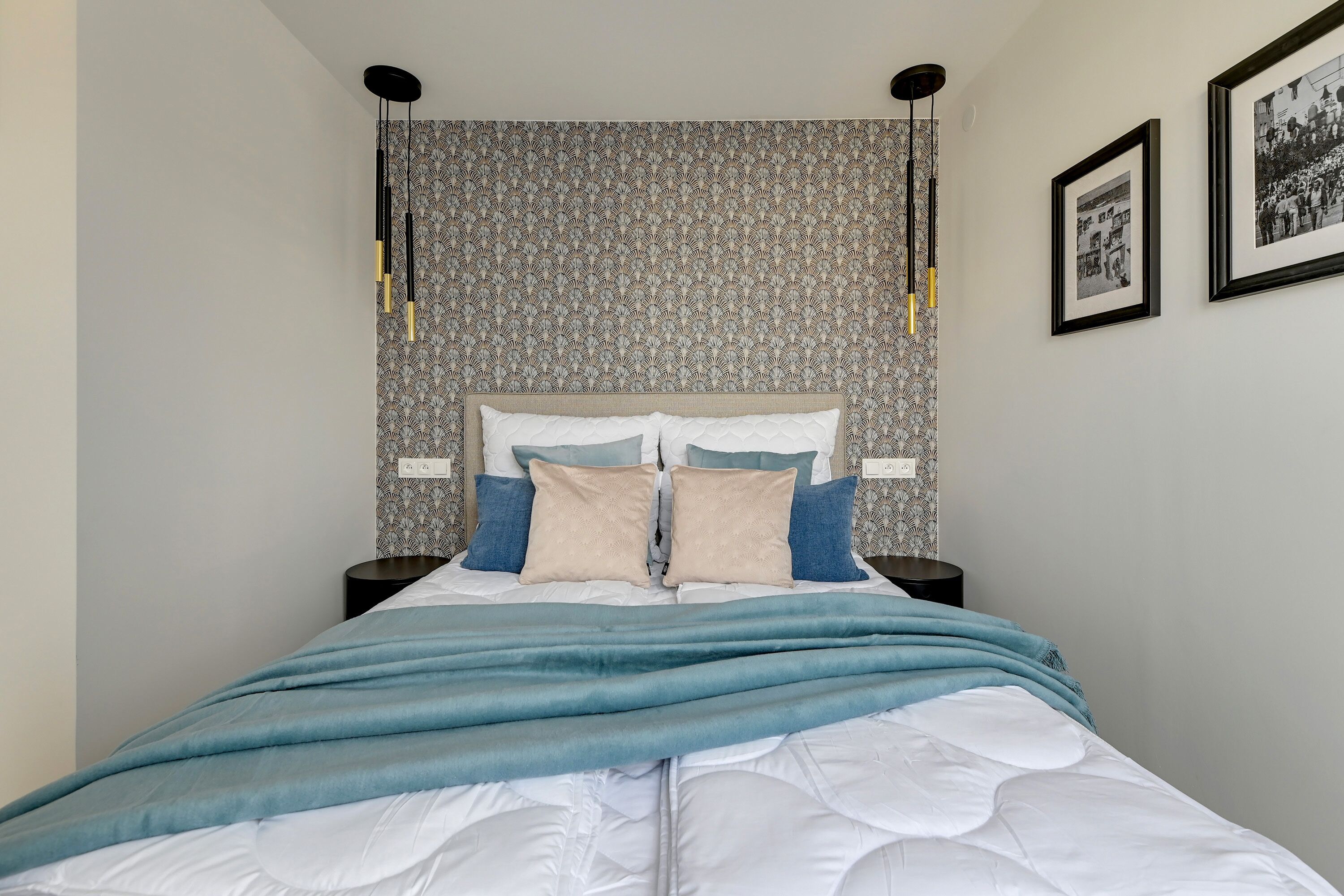 A neatly made bed with a blue blanket, a patterned wallpaper, and two black wall-mounted lamps.