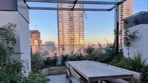 Een terras op het dak, met een houten tafel en banken, omgeven door groen en met uitzicht op een stadsgezicht met hoge gebouwen.