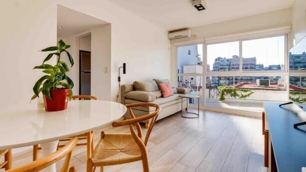 Uma sala de estar moderna com uma mesa de jantar redonda, cadeiras de madeira, um sofá e uma grande janela com vista para a cidade.