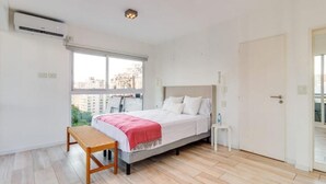 Quarto com cama, mesa de cabeceira, um banco e janela com vista para edifícios.