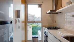 Cozinha estreita com armários brancos, fogão e janela com vista para edifícios e vegetação.
