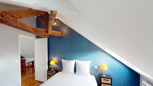 Une chambre avec un plafond en pente, des poutres en bois, un lit recouvert de draps blancs, deux tables de chevet avec des lampes et un fauteuil rouge dans un coin.