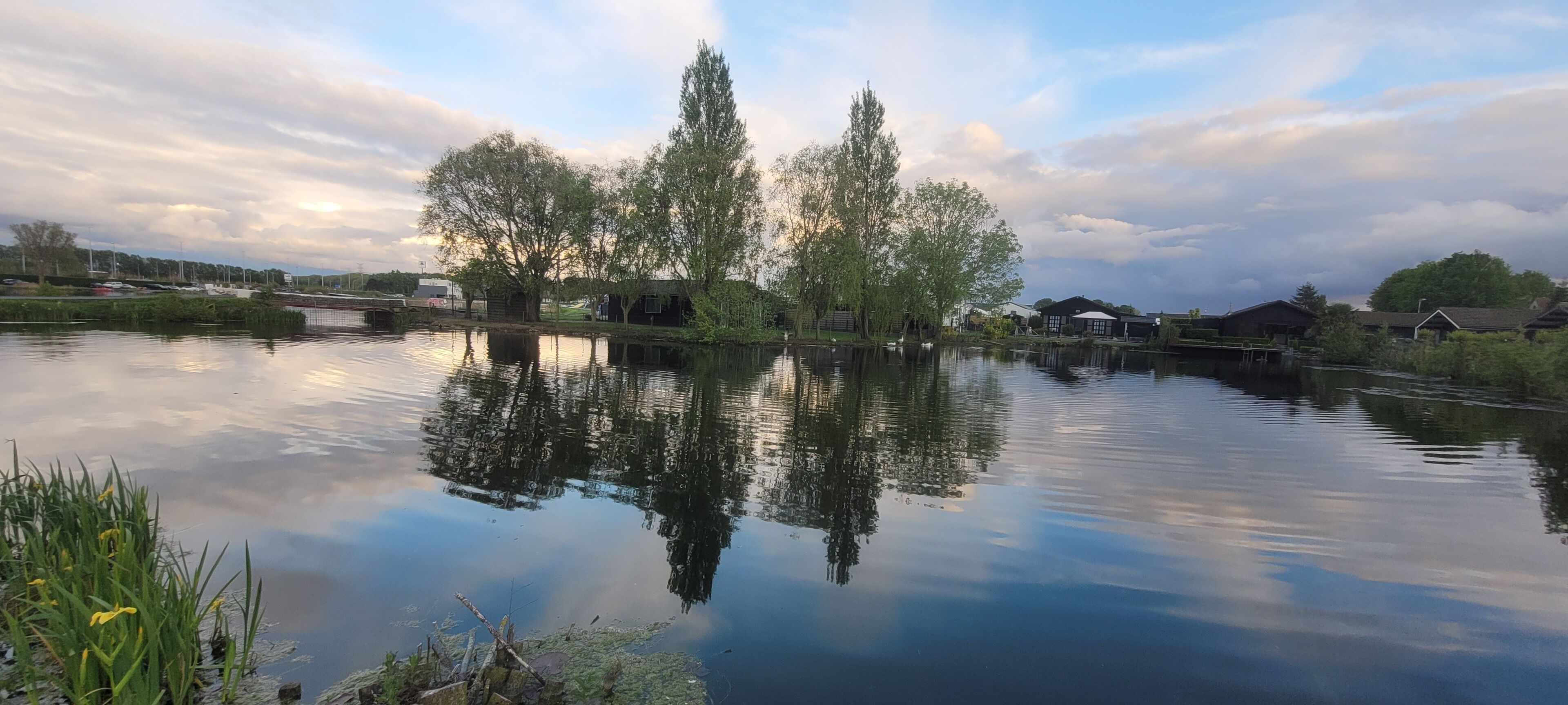 A calm river with reflections of trees and buildings, surrounded by greenery and a clear sky.