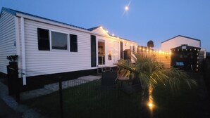 A mobile home with a fenced yard, a small patio, and string lights. - Chalet Sea-renity Lakeside met Jacuzzi (Jabekke)