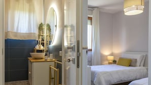 Ein modernes Hotelzimmer mit Bad, einem Bett mit weißer Bettwäsche, einem Nachttisch mit Lampe und einer kleinen Sitzecke mit Pflanze.