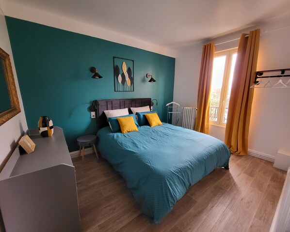 A bedroom with a bed, a dresser, a nightstand, a window with curtains, and a wall with a framed picture.