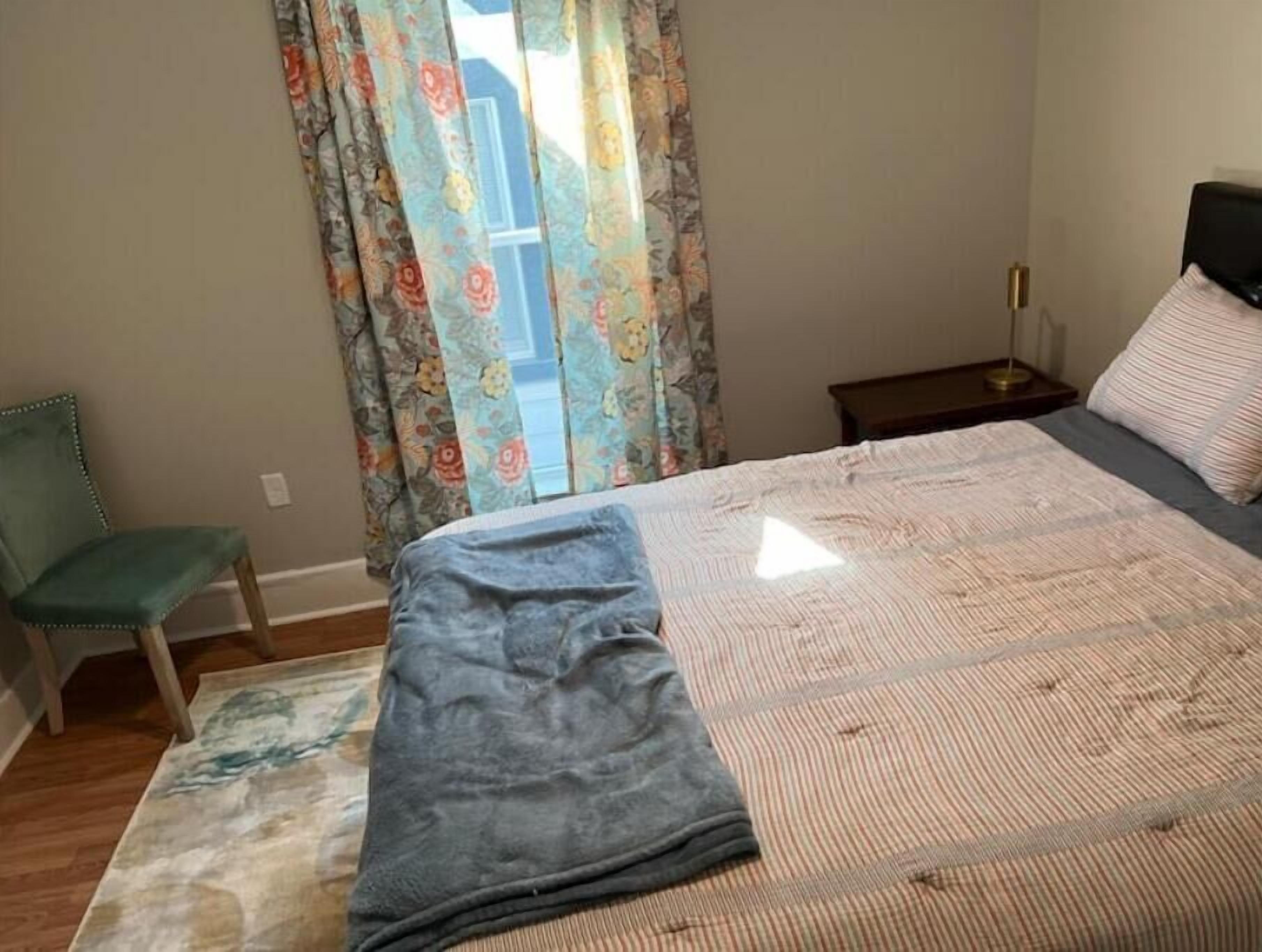 A bedroom with a bed, a chair, a nightstand, and a window with floral curtains.