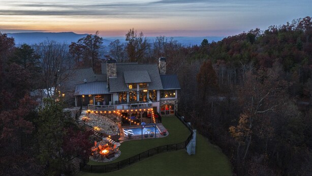 Una casa grande con piscina e iluminación exterior, rodeada de árboles y vistas a las montañas.