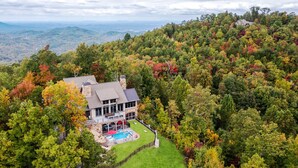 Una casa grande con piscina y terraza, rodeada de un bosque denso y montañas al fondo.