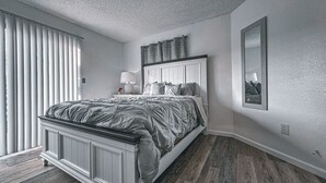 Ein Schlafzimmer mit Bett, Nachttisch, Lampe, Spiegel und Fenster mit Jalousien.