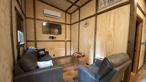 Interior acogedor de una cabaña con dos sofás de cuero, una pared de madera con un letrero que dice 'Welcome Home Cabin', un televisor montado y una mesita con una cesta.