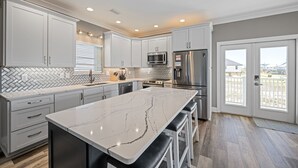 A modern kitchen with white cabinets, a marble island, stainless steel appliances, and a view to the outside. - 5BR Beach House+Steps To Beach+Balcony Gulf Views+Firepit+Elevator+Dog Friendly (Gulf Shores)