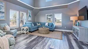 A spacious living room with a blue sofa, a round coffee table, and a large window with blinds. - 5BR Beach House+Steps To Beach+Balcony Gulf Views+Firepit+Elevator+Dog Friendly (Gulf Shores)