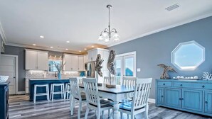 A modern kitchen with white cabinets, a blue island, and a dining area with white chairs. - 5BR Beach House+Steps To Beach+Balcony Gulf Views+Firepit+Elevator+Dog Friendly (Gulf Shores)