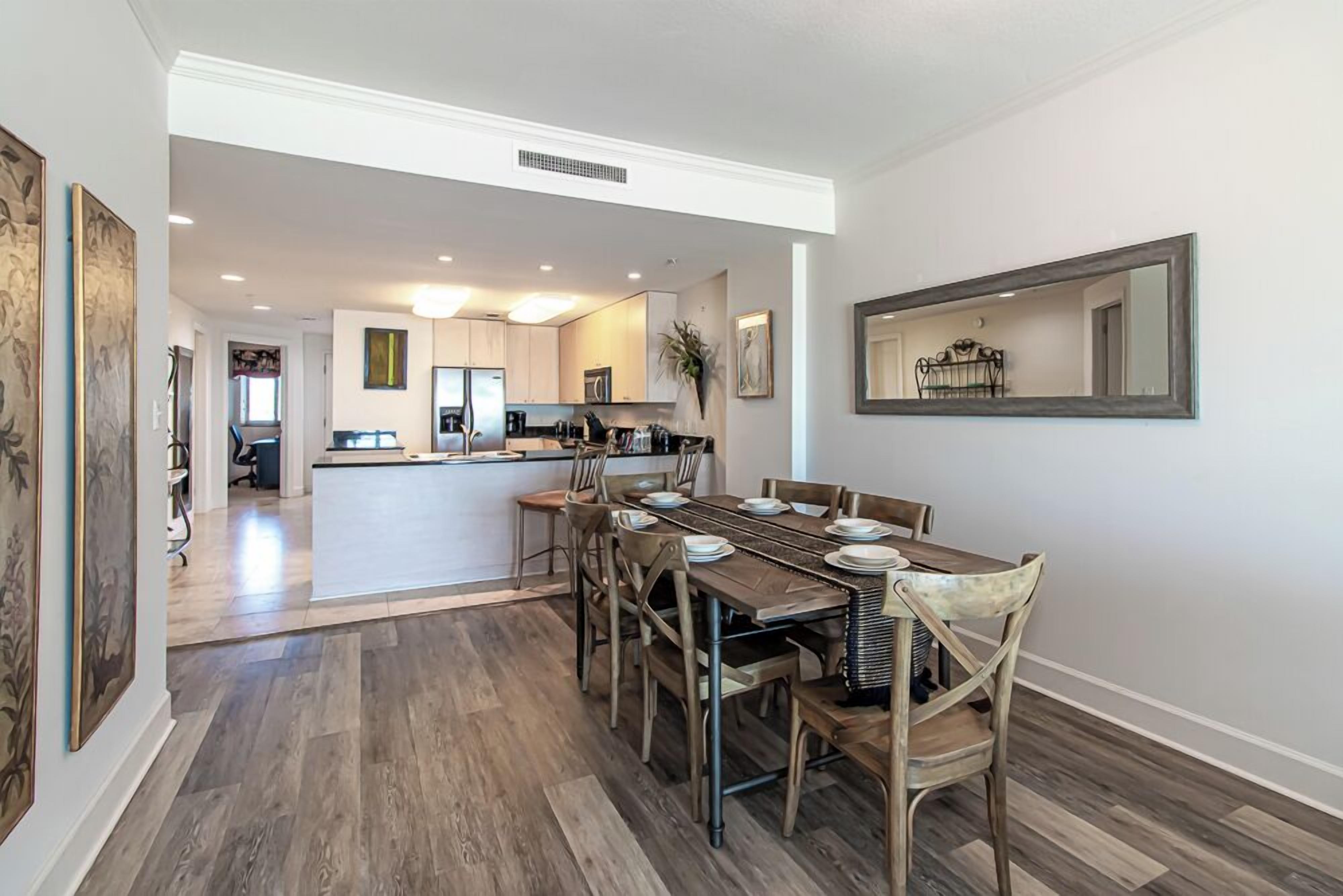 A spacious dining area with a long wooden table and chairs, a kitchen in the background, and a large mirror on the wall.
