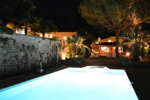 A swimming pool at night with a stone wall and a building in the background.