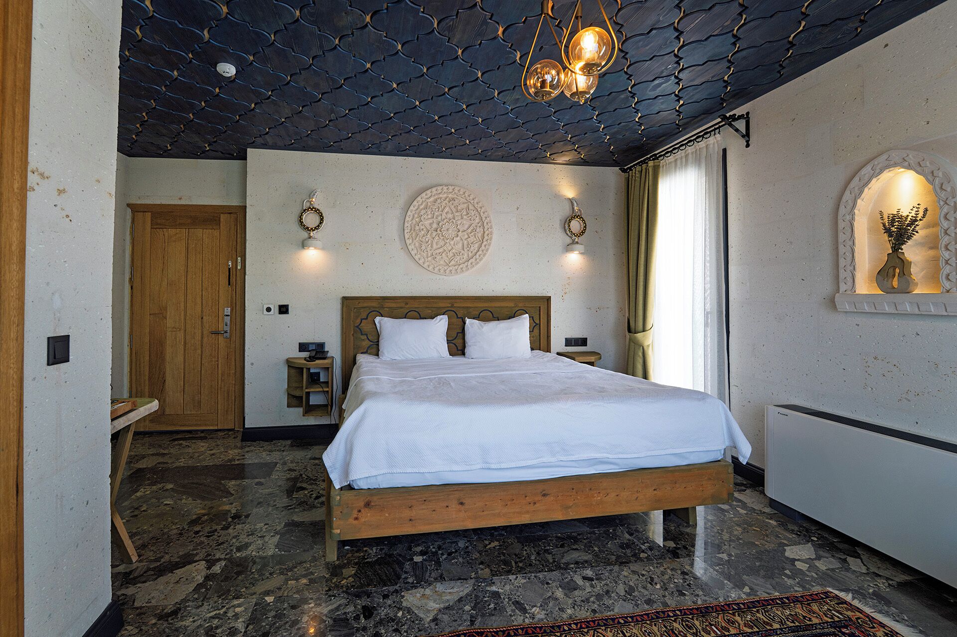 A bedroom with a wooden bed, a stone floor, a wooden door, and a ceiling with a unique pattern.