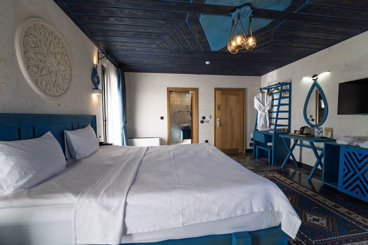 A bedroom with a large bed, a wooden ceiling, a blue chair, a desk, and a mirror.