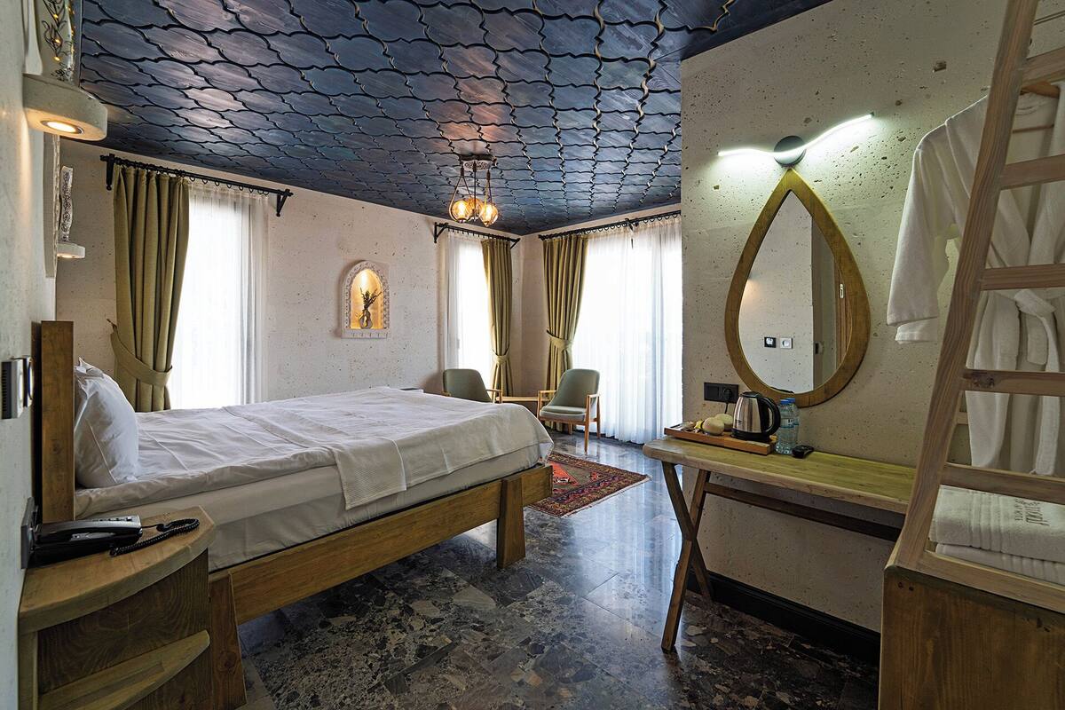 A hotel room with a bed, a desk with a mirror, a chair, a small table with a kettle, and a window with curtains.