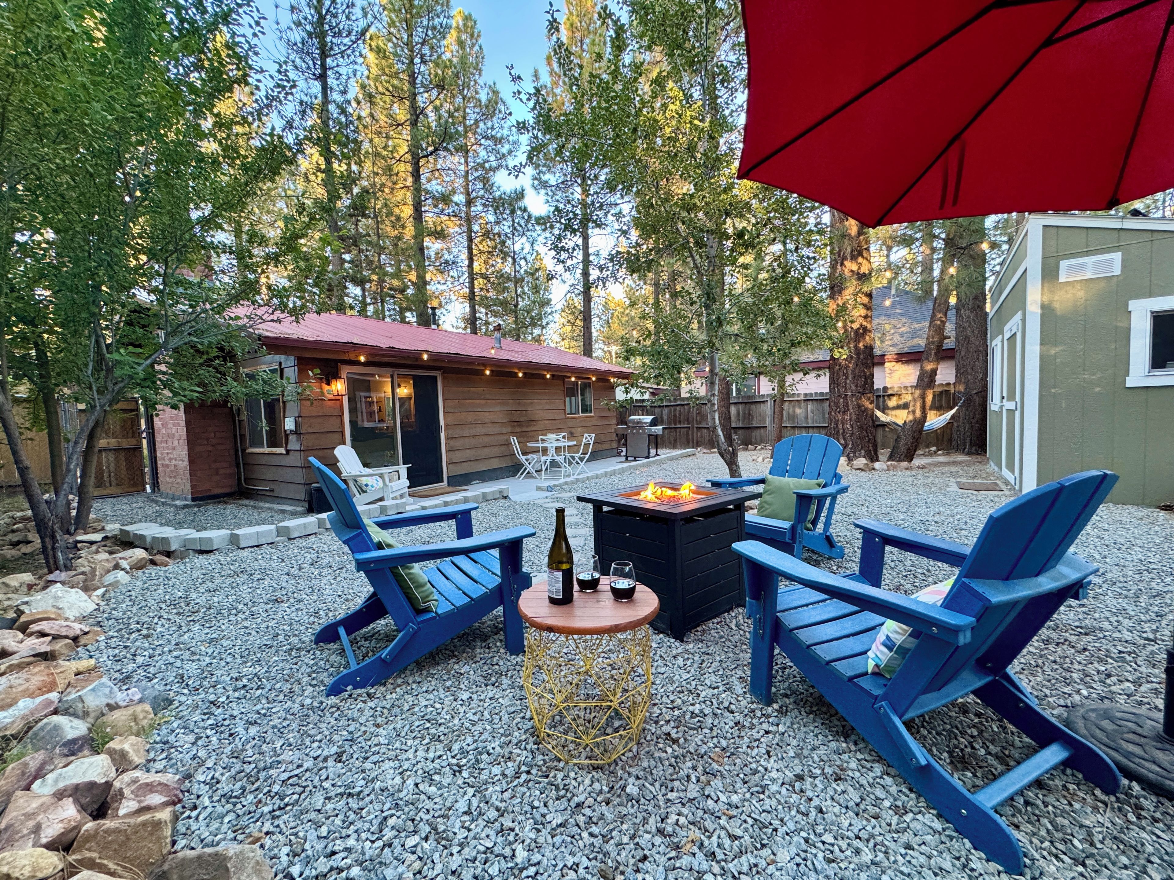 An outdoor patio with blue Adirondack chairs, a fire pit, and a small table with a bottle and glasses.