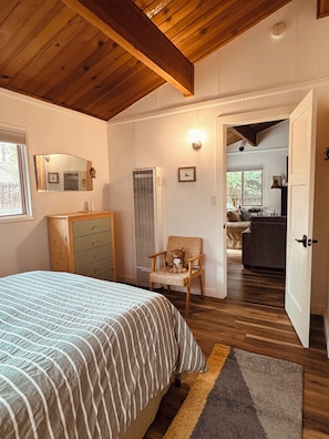 A bedroom with a bed, dresser, chair, and a hallway leading to a living area.