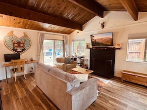 A cozy living room with a fireplace, wooden flooring, a sofa, and a desk with a computer.