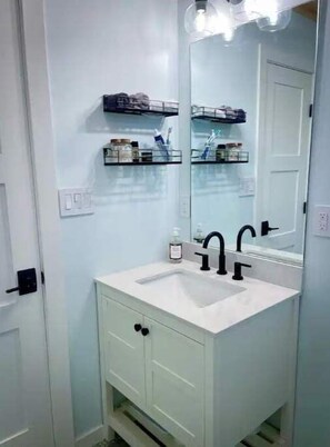 Un baño con tocador blanco, grifos negros y un espejo grande. Hay dos repisas sobre el lavamanos con artículos de aseo.