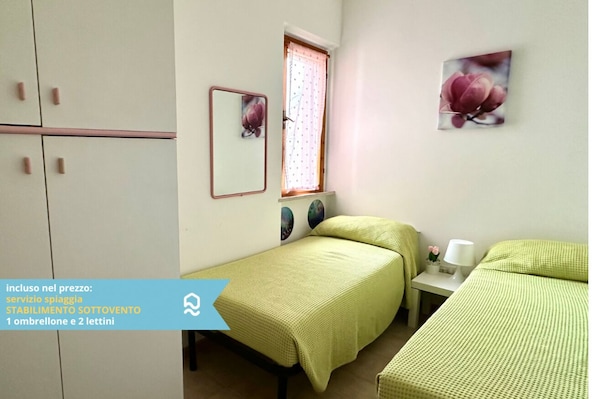 A bedroom with two beds, a window with a curtain, a bedside table with a lamp, and a wall-mounted mirror.