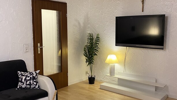 En moderne stue med en sort sofa, et rundt sofabord, en potteplante, et fladskærms-tv monteret på væggen og et træloft.