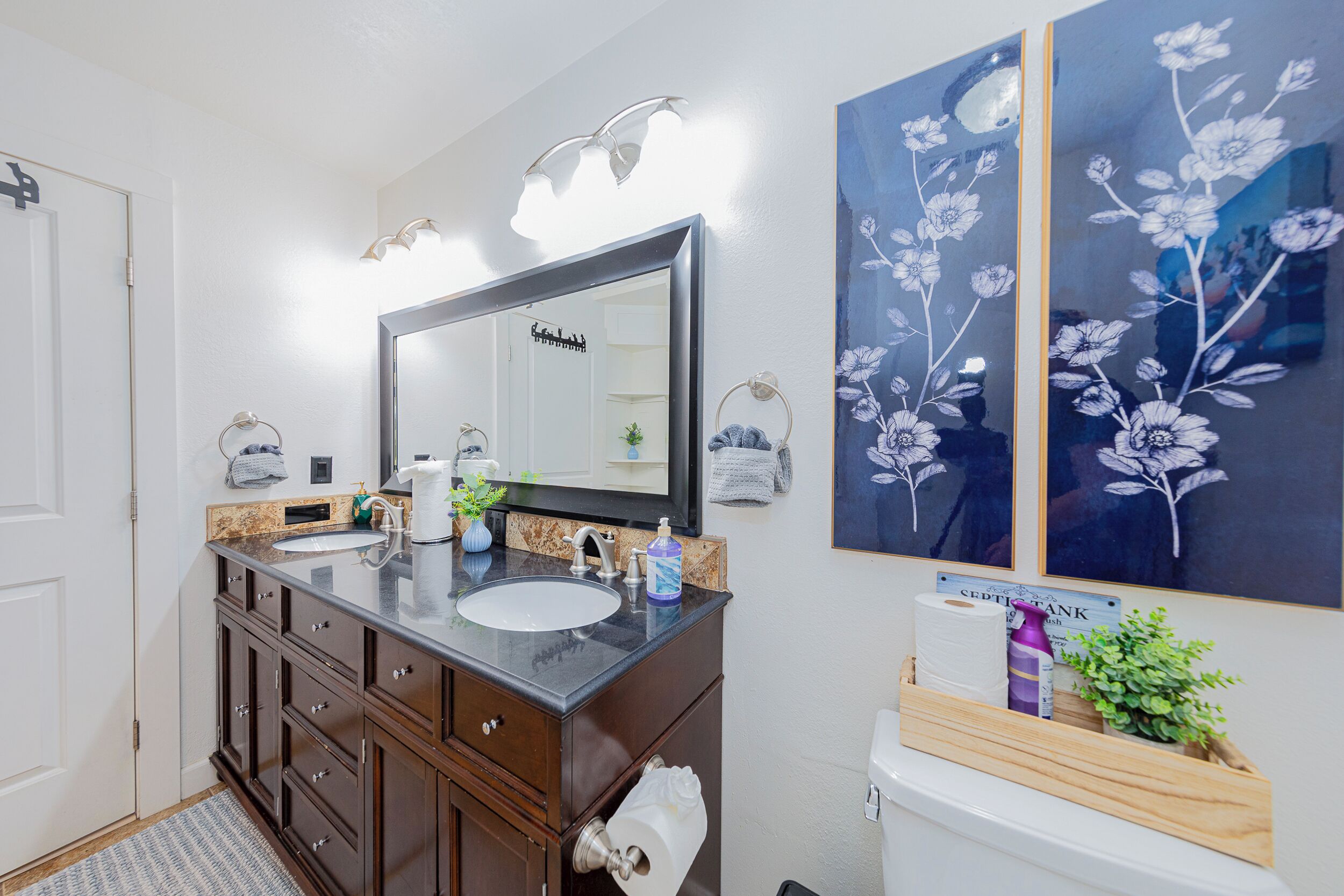 Un baño con tocador de madera oscura, un lavamanos blanco, un espejo grande y dos cuadros enmarcados con motivos florales en la pared.