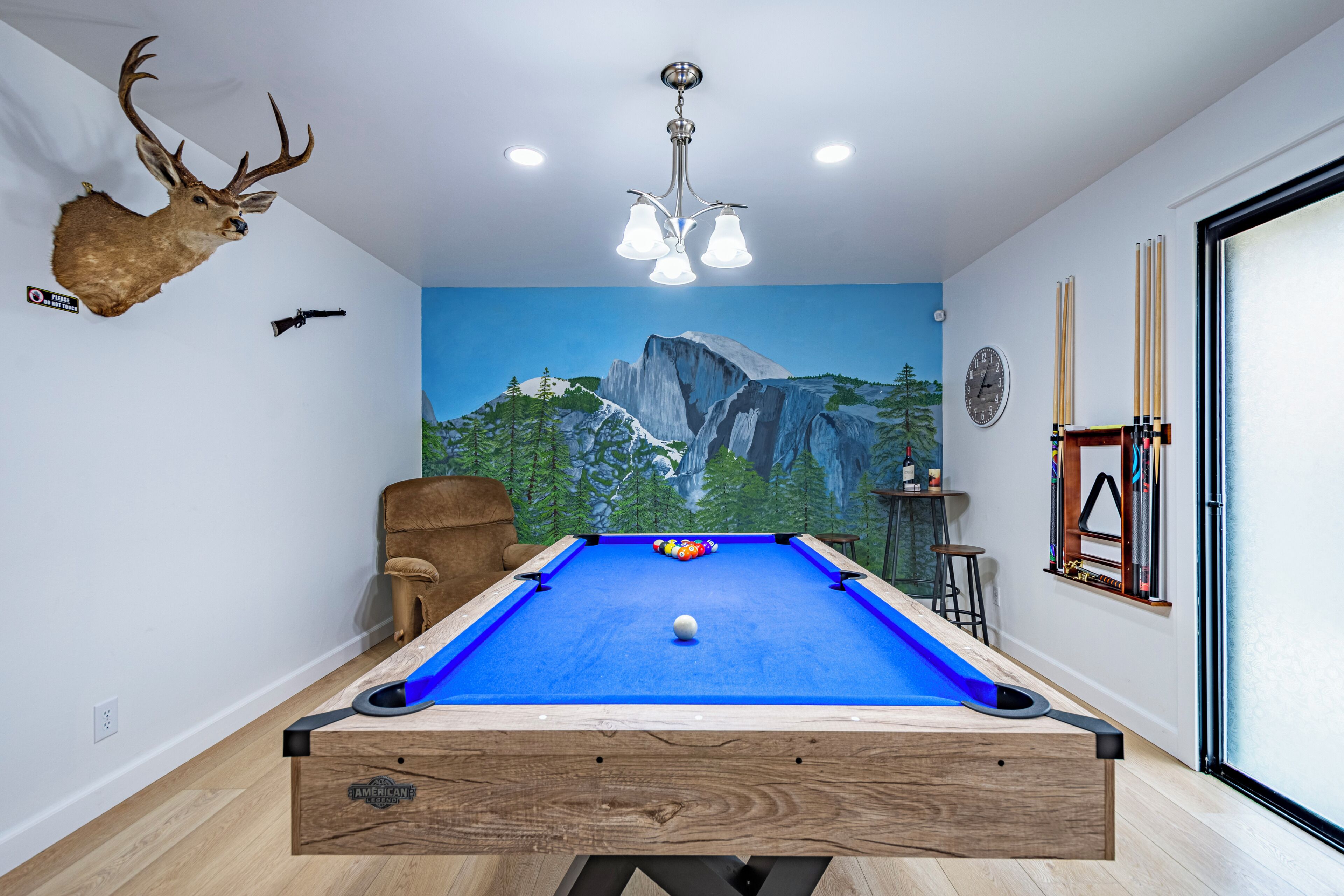 A pool table with a blue felt surface, a mounted deer head, and a mural of a mountain landscape.