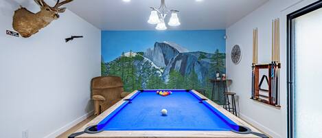 A pool table with a blue felt surface, a mounted deer head, and a mural of a mountain landscape.