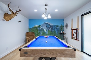 Una mesa de billar con superficie de fieltro azul, una cabeza de ciervo montada en la pared y un mural con paisaje de montaña.