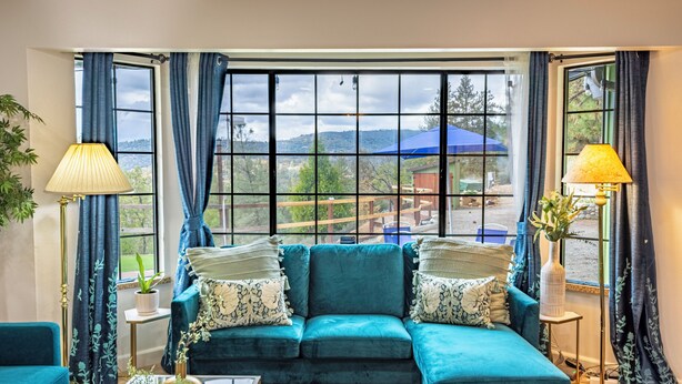 A room with a teal sofa, a coffee table, a lamp, and a view of a blue umbrella and greenery outside.