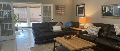 A living room with a black leather sofa, a wooden coffee table, and a lamp. There is a door leading to a patio area with a view of the outside.