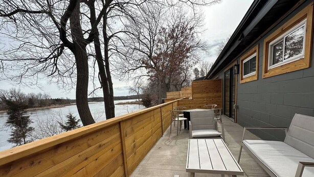 Una terraza de madera con vistas a un lago, mobiliario exterior y un edificio con ventanas.
