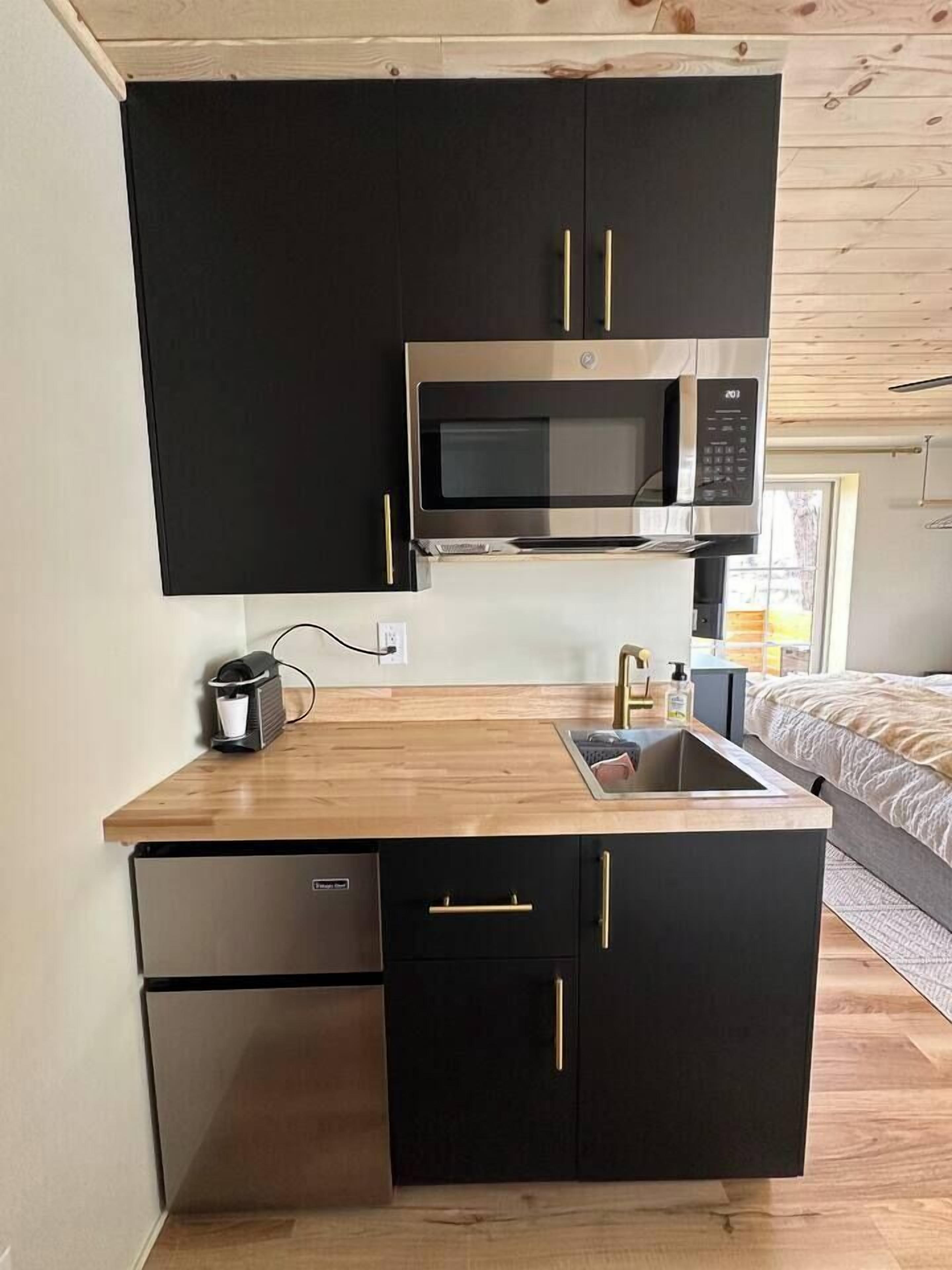 A compact kitchen with black cabinets, a stainless steel microwave, a coffee machine, and a small refrigerator.