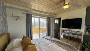 A bedroom with a large bed, a ceiling fan, a TV, and a sliding door to a balcony. - Nordic Harbor | Room 5 | Cedar Shadows (Center City)