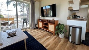 Un salon moderne avec un plancher en bois, un téléviseur fixé au mur, une table à manger avec un panier, une plante et une poubelle.