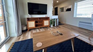 Un salon doté d’un plancher en bois, d’un grand téléviseur, d’un meuble en bois, d’une petite table et d’une fenêtre avec des stores.