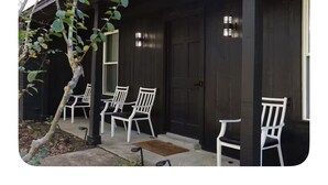 Una casa in legno scuro con un portico, tre sedie bianche e un albero.