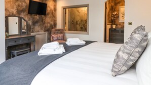 Habitación de hotel con una cama grande, una zona de estar con una silla y una mesa, un televisor de pantalla plana en la pared y una ventana con vistas al exterior.