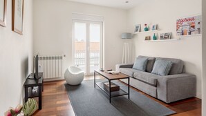 Una sala de estar moderna con un sofá gris, una mesita de centro y una silla blanca. Se observa un radiador, una lámpara y una estantería con fotografías y jarrones.