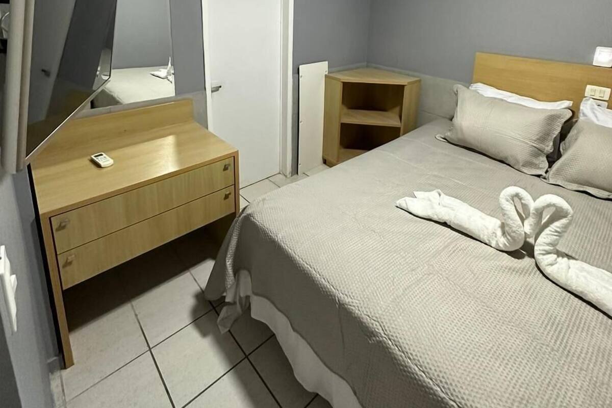 A hotel room with a bed, a bedside table, a mirror, a wardrobe, and an air conditioning unit.