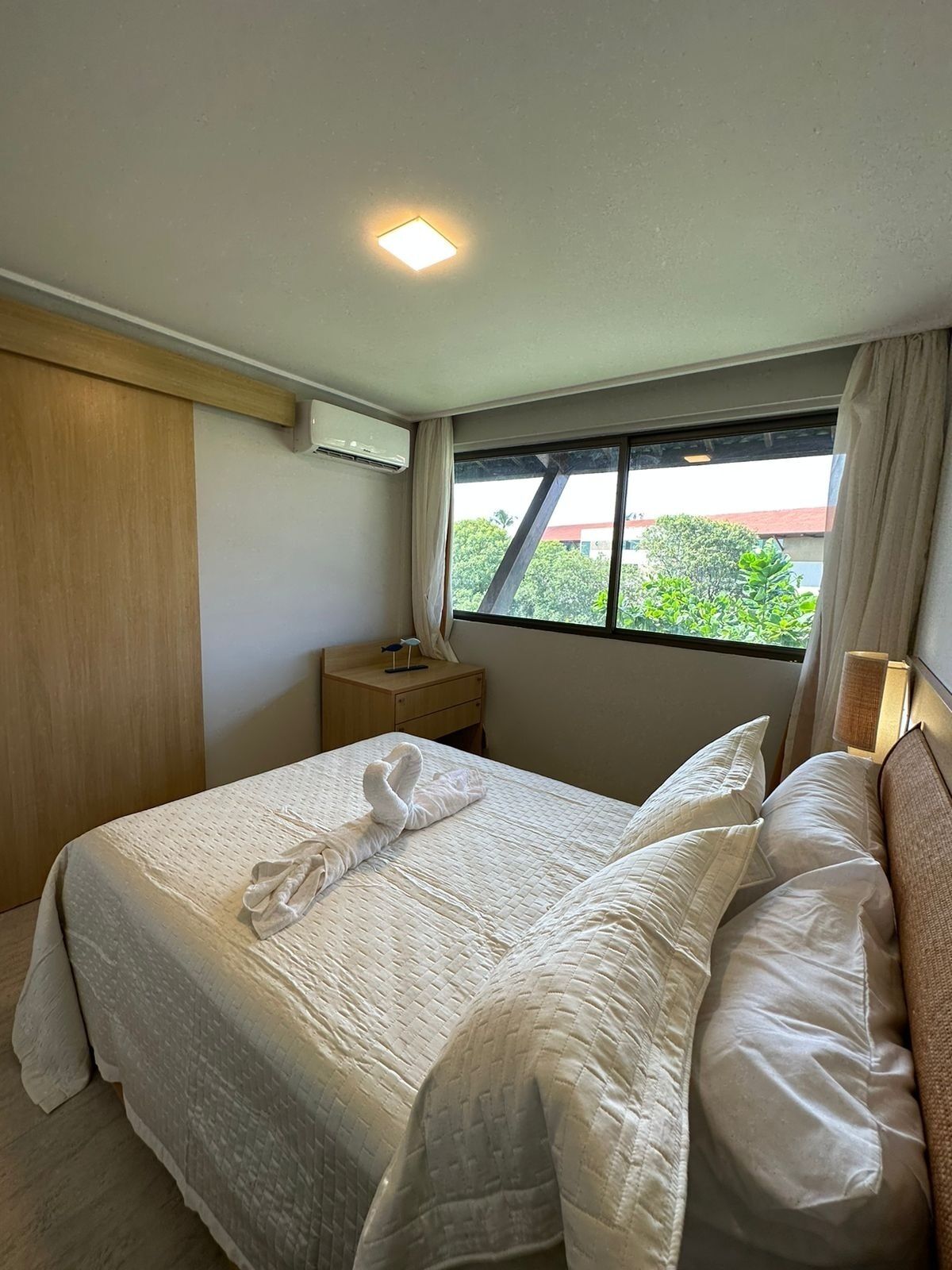 Una habitación de hotel con cama, mesita de noche, equipo de aire acondicionado, ventana con vistas a zonas verdes y una lámpara en el techo.