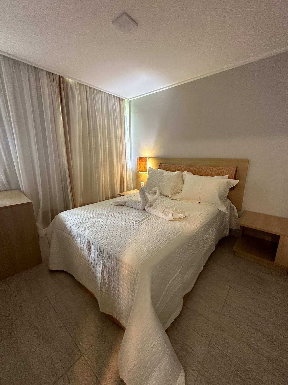 Uma cama bem arrumada com um edredom branco, uma cabeceira de madeira e uma mesinha de cabeceira.