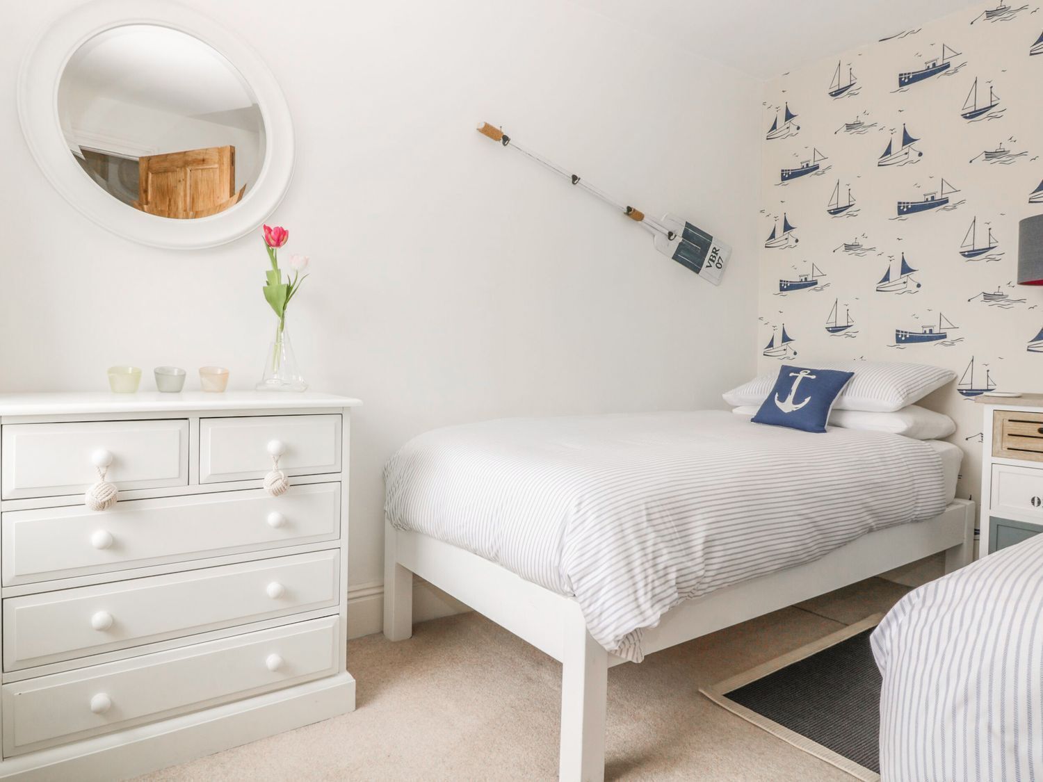 Un dormitorio con cama blanca, cómoda con cajones, espejo redondo, un florero con flores y papel tapiz con motivos náuticos.