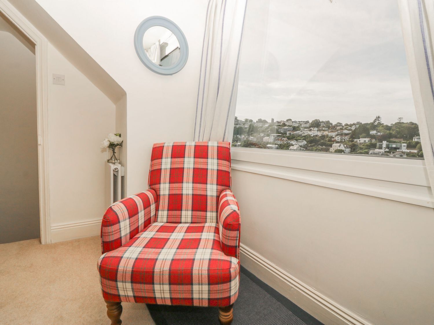 A corner room with a red plaid armchair, a round mirror, and a window with a city view.